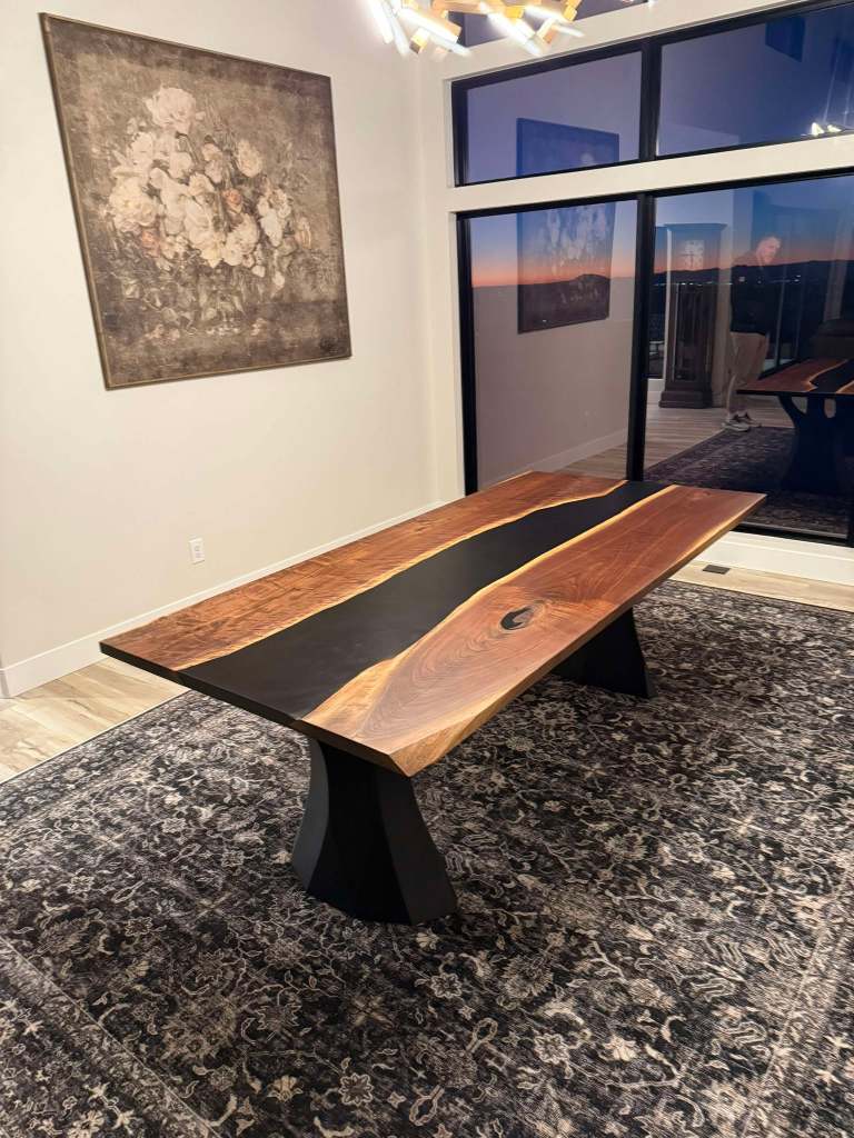 A handcrafted black walnut dining table featuring a live-edge design and modern polished steel legs, set in an elegantly decorated dining room with a rug and artwork on the walls.