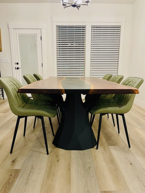 A handmade dining table crafted from black walnut with live edges and black epoxy resin, featuring a modern brushed steel base and surrounded by green chairs.