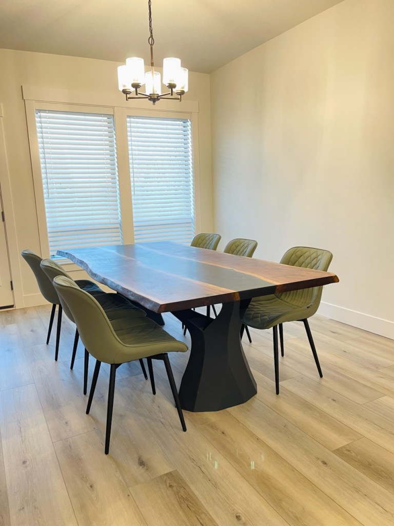 A handcrafted dining table made from black walnut slabs with a black epoxy resin finish, surrounded by modern green chairs in a well-lit room with white walls and large windows.