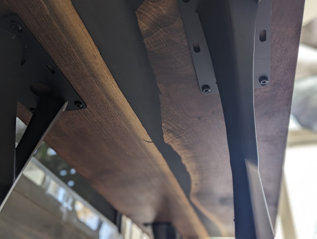 Close-up view of the underside of a handcrafted black walnut dining table, featuring live-edge detail and modern polished steel legs, showcasing the craftsmanship and design.