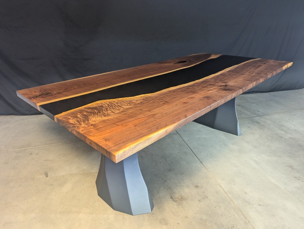A handcrafted black walnut dining table with a modern design featuring polished steel legs and a live-edge detail, placed on a concrete floor against a dark backdrop.