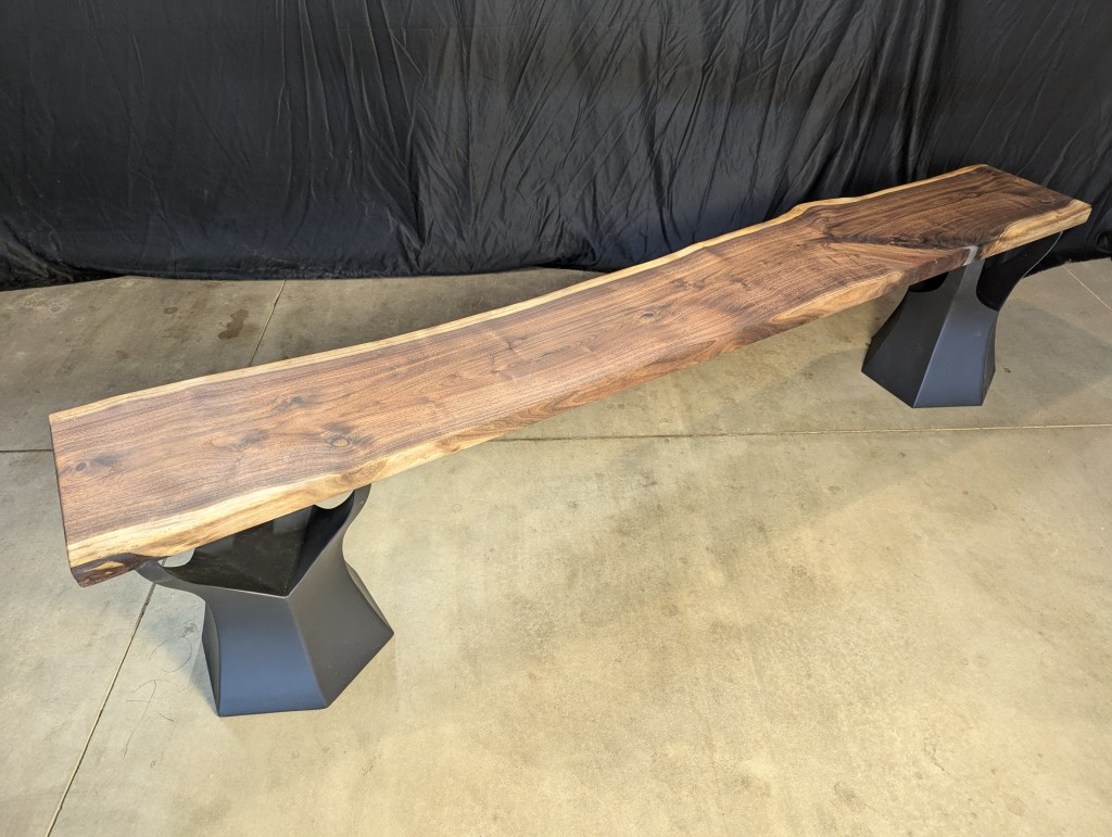 A handcrafted wooden bench with a natural edge top and black metal legs, displayed against a dark background.