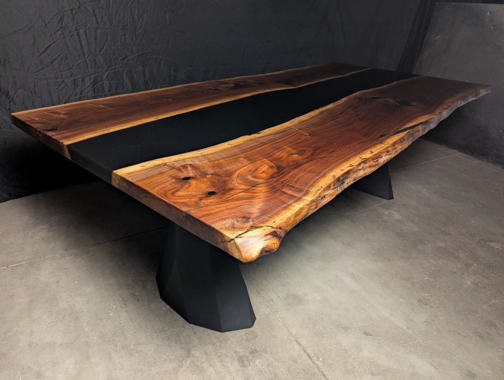A handmade black walnut dining table featuring a black epoxy resin stripe, with live edges. The table has brushed steel legs and dimensions of 90" in length and 42" to 46" in width.