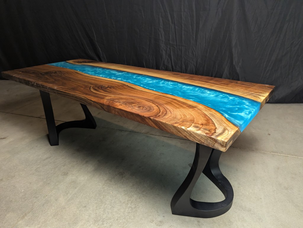 A handcrafted black walnut river table featuring a Caribbean blue epoxy river, supported by brushed steel legs, showcasing live edges and unique grain character.