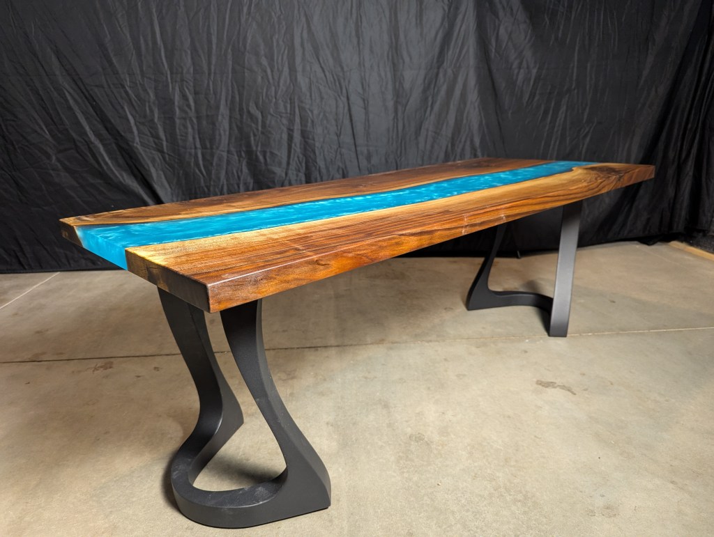 A handcrafted black walnut river table featuring a vibrant blue epoxy river running through the center, supported by brushed steel legs, showcased against a black backdrop.