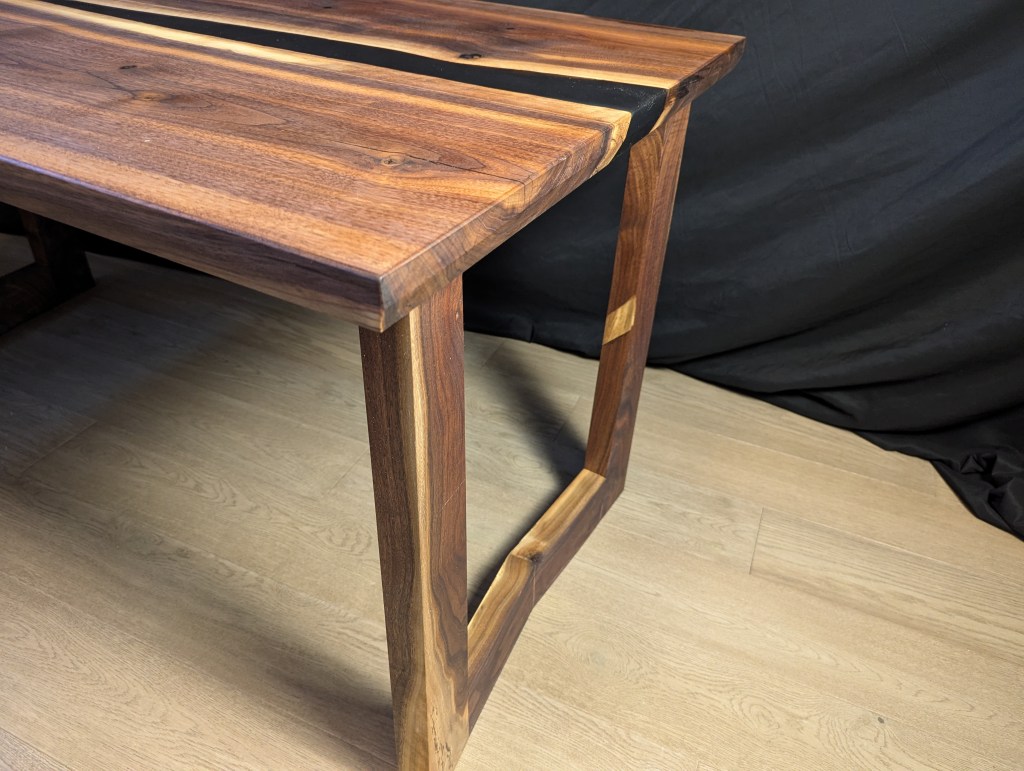A close-up view of a handcrafted Highline Executive Desk made from solid black walnut, showcasing the sleek live edge top and angular legs, highlighted by a matte black epoxy river.